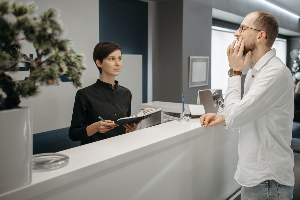 A female reception dental nurse at a dental clinic holding a book and looking at a patient explaining his toothache